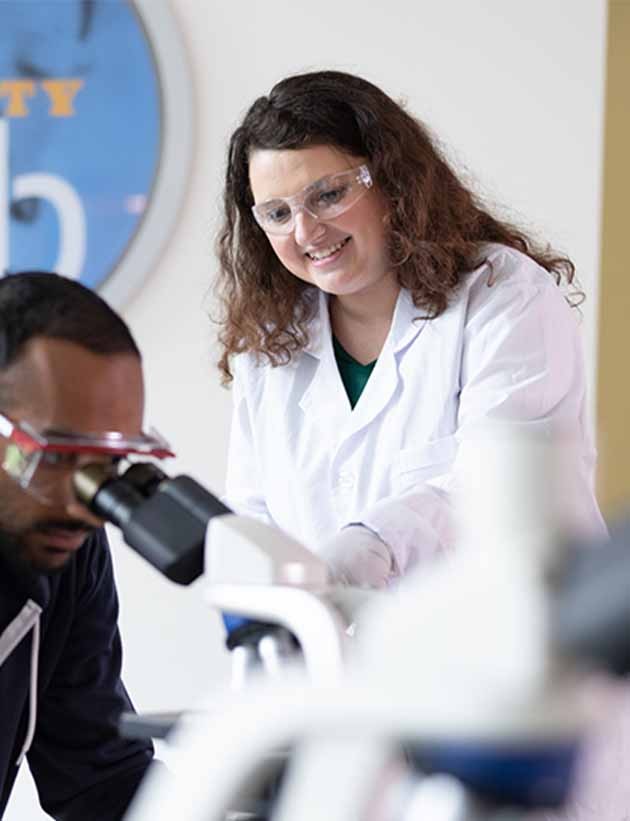 two students looking into microscopes with teacher in lab coat smiling next to them