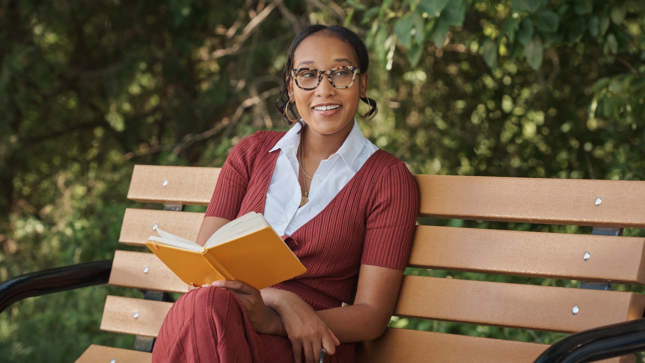 Lauren, living with MS, sitting on a bench outside smiling and holding a book