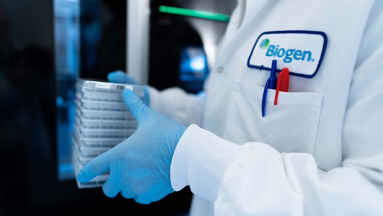 Close up of Biogen employee in a Biogen lab coat with pens in it, wearing blue gloves while working in the lab